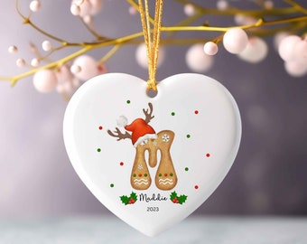 Può includere: Ornamento natalizio bianco a forma di cuore con un design a renna di pan di zenzero. La renna indossa un cappello da Babbo Natale e ha il nome "Maddie" e l'anno "2023" stampati sopra. Appeso a un nastro dorato.