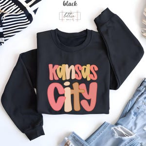 以下が含まれることがあります： レトロなフォントで「Kansas City」の文字が入った黒いクルーネックのスウェットシャツ。テキストはピンク、黄色、茶色の色合いです。スウェットシャツは、ジーンズと一緒に白い表面に表示されています。
