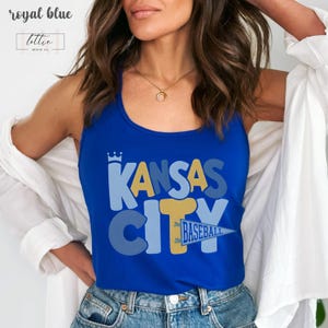 Op de afbeelding: Koningsblauwe tanktop met de tekst "KANSAS CITY BASEBALL" in een gelaagd ontwerp. De top heeft een kroongrafiek boven het woord "KANSAS". De tanktop wordt gedragen met een spijkerbroek en een wit overhemd.