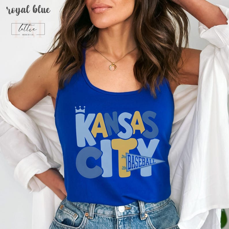 Op de afbeelding: Koningsblauwe tanktop met de tekst "KANSAS CITY BASEBALL" in een gelaagd ontwerp. De top heeft een kroongrafiek boven het woord "KANSAS". De tanktop wordt gedragen met een spijkerbroek en een wit overhemd.