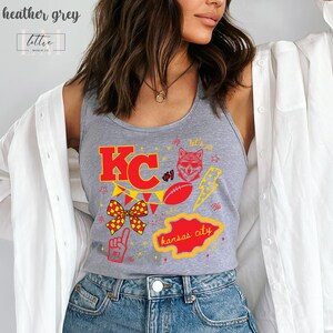 May include: Heather grey tank top with a red and yellow graphic design. The design includes the letters "KC", a football, a lightning bolt, a fox head, and the words "Kansas City". The tank top also has the text "#1".