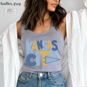 Op de afbeelding: Heather grijze tanktop met de tekst "KANSAS CITY" in blauwe en gele blokletters. De tanktop heeft een ronde hals en is gemaakt van een zacht materiaal. De top maakt deel uit van een kledingcollectie.