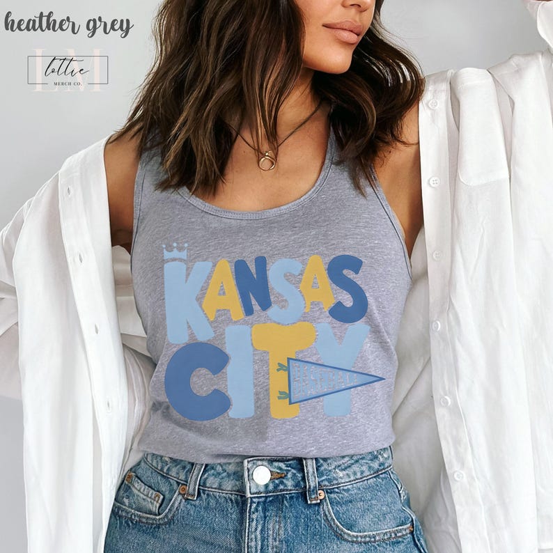 Op de afbeelding: Heather grijze tanktop met de tekst "KANSAS CITY" in blauwe en gele blokletters. De tanktop heeft een ronde hals en is gemaakt van een zacht materiaal. De top maakt deel uit van een kledingcollectie.