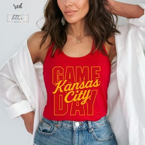 May include: Red tank top with the text "GAME Kansas City DAY" in yellow lettering. The tank top is worn with blue jeans and a white shirt. The top has the word "red" and the brand name "lottie merch co" in the upper left corner.