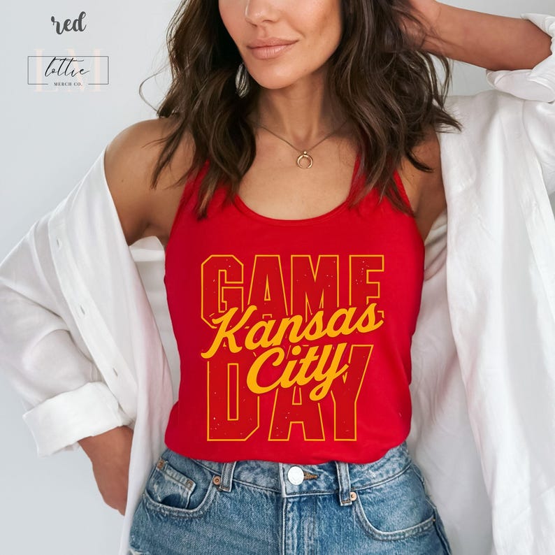 May include: Red tank top with the text "GAME Kansas City DAY" in yellow lettering. The tank top is worn with blue jeans and a white shirt. The top has the word "red" and the brand name "lottie merch co" in the upper left corner.