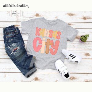 以下が含まれることがあります： カラフルなレトロフォントで「Kansas City」という文字がプリントされたグレーのTシャツ。青いジーンズと黒いストライプの白いスニーカーと組み合わせています。「athletic heather」という文字が左上にあります。