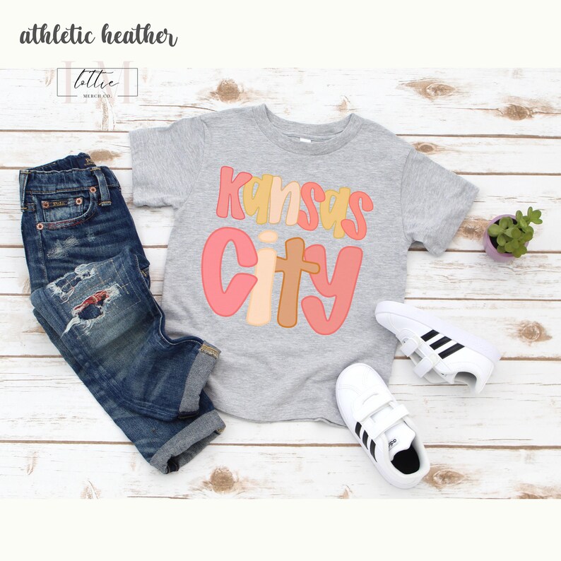 以下が含まれることがあります： カラフルなレトロフォントで「Kansas City」という文字がプリントされたグレーのTシャツ。青いジーンズと黒いストライプの白いスニーカーと組み合わせています。「athletic heather」という文字が左上にあります。