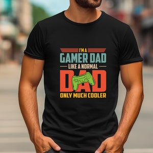 May include: Black t-shirt with the text "I'M A GAMER DAD LIKE A NORMAL DAD ONLY MUCH COOLER" in teal, orange, and yellow. A green video game controller graphic is in the centre of the word "DAD".