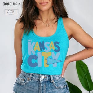Op de afbeelding: Een turquoise tanktop met de woorden "KANSAS CITY" in een kleurrijk, speels lettertype. Het woord "CITY" wordt gedeeltelijk bedekt door een wimpel met de tekst "BASEBALL". De top is gelabeld "tahiti blue" en heeft een klein logo.