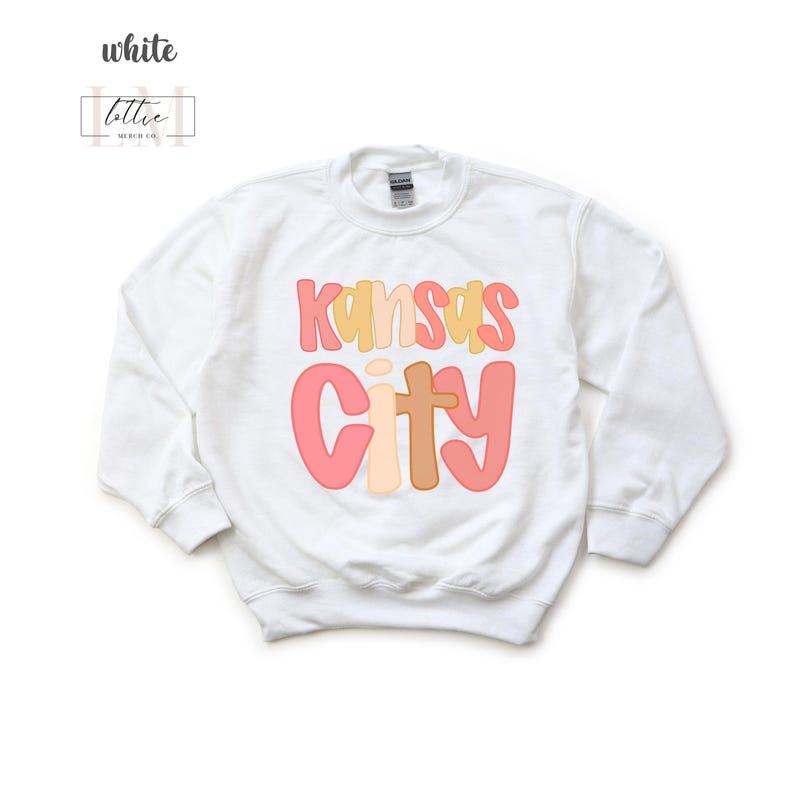 以下が含まれることがあります： カラフルなレトロフォントで「Kansas City」と書かれた白いスウェットシャツ。「Kansas」は黄色とピンク、「City」はピンクと茶色です。クルーネックと長袖のデザインです。