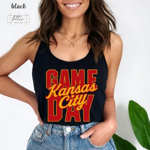 May include: Black tank top with the words "GAME Kansas City DAY" in red and yellow lettering. The tank top is worn by a person in blue jeans. The top has a racerback design.
