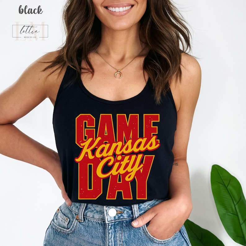 May include: Black tank top with the words "GAME Kansas City DAY" in red and yellow lettering. The tank top is worn by a person in blue jeans. The top has a racerback design.