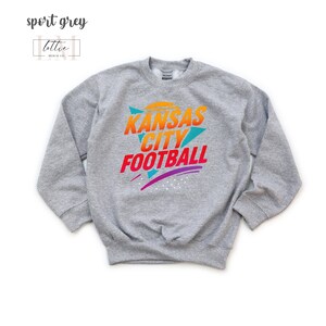 K&ouml;nnte beinhalten: Sportgraues Sweatshirt mit dem Text "Kansas City Football" in Orange, Gelb und Rot. Das Sweatshirt hat ein Retro-Design mit t&uuml;rkisfarbenen und lila Akzenten. Der Markenname "lottie merch co." befindet sich in der oberen linken Ecke.