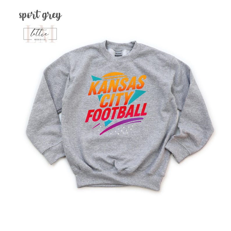 K&ouml;nnte beinhalten: Sportgraues Sweatshirt mit dem Text "Kansas City Football" in Orange, Gelb und Rot. Das Sweatshirt hat ein Retro-Design mit t&uuml;rkisfarbenen und lila Akzenten. Der Markenname "lottie merch co." befindet sich in der oberen linken Ecke.