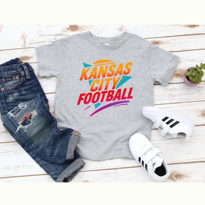 K&ouml;nnte beinhalten: Graues T-Shirt mit dem Schriftzug "Kansas City Football" in einem farbenfrohen Retro-Design. Das Bild zeigt auch blaue Jeans, wei&szlig;e Turnschuhe mit schwarzen Streifen und eine kleine Topfpflanze.