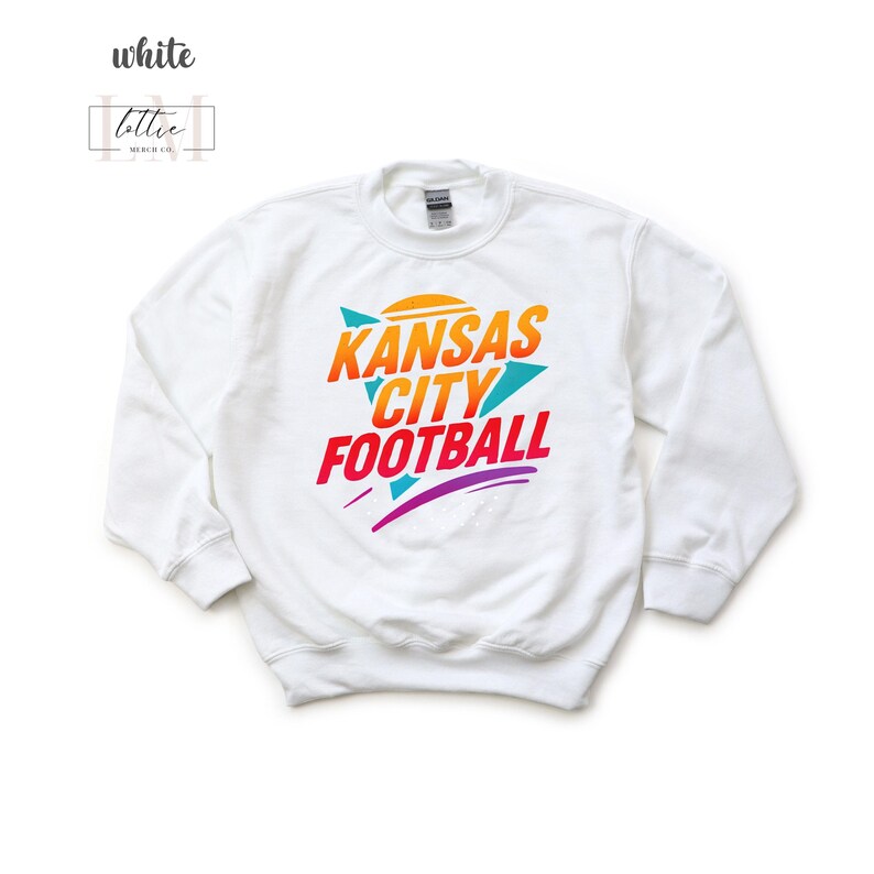K&ouml;nnte beinhalten: Wei&szlig;es Sweatshirt mit dem Text "Kansas City Football" in Orange, T&uuml;rkis und Lila. Das Design beinhaltet eine stilisierte Sonne und Pfeilformen. Das Sweatshirt ist von der Marke Gildan.