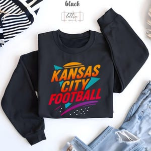 K&ouml;nnte beinhalten: Schwarzes Sweatshirt mit Rundhalsausschnitt und dem Text "Kansas City Football" in einem Retro-Design. Der Text ist in Orange, Rot, T&uuml;rkis und Lila gehalten. Das Wort "black" befindet sich oben links im Bild.