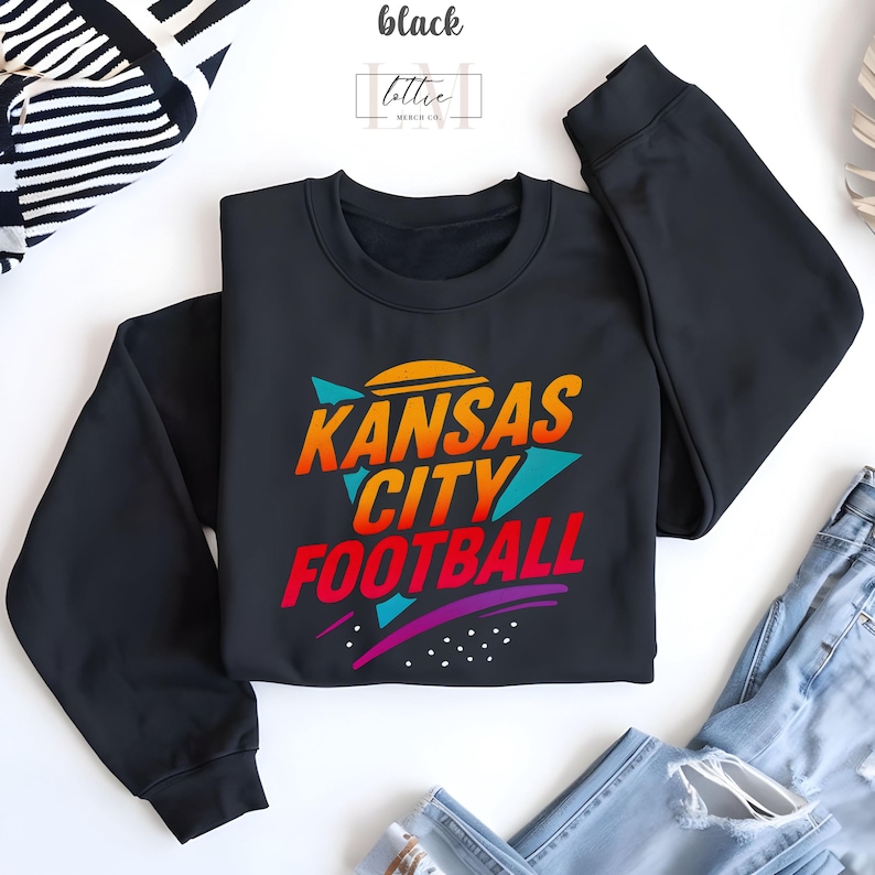 K&ouml;nnte beinhalten: Schwarzes Sweatshirt mit Rundhalsausschnitt und dem Text "Kansas City Football" in einem Retro-Design. Der Text ist in Orange, Rot, T&uuml;rkis und Lila gehalten. Das Wort "black" befindet sich oben links im Bild.