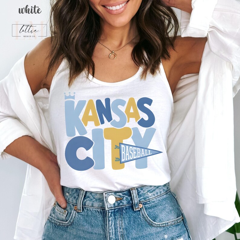 Op de afbeelding: Witte tanktop met de tekst "KANSAS CITY BASEBALL" in blauwe en gouden letters. Het woord "Kansas" heeft er een kroon boven. De tanktop is een casual stijl.