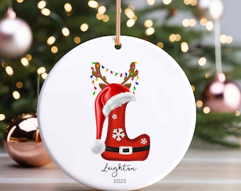 Può includere: Addobbo natalizio in ceramica bianca con un calzino rosso con un cappello di Babbo Natale in cima. Il calzino ha un fiocco di neve bianco e una cintura nera. L'addobbo è decorato con luci colorate e corna di renna. Il testo "Leighton 2023" è stampato sull'addobbo.