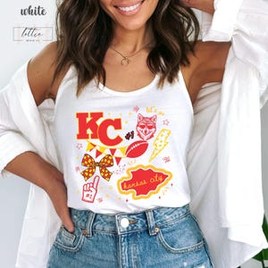 May include: White tank top with a Kansas City design. The design includes the letters "KC" in red, a football, a wolf wearing sunglasses, and the words "Kansas City". The tank top is a fan item.