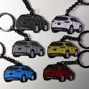 May include: A collection of car-shaped keychains in various colors, including black, white, gray, yellow, blue, and red. Each keychain features a detailed car design with black chain and a silver key ring.