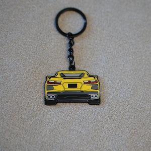 May include: A yellow car-shaped keyring with a black border and accents. The keyring has a black key ring and chain. The car's rear design is visible, including taillights and exhaust pipes.