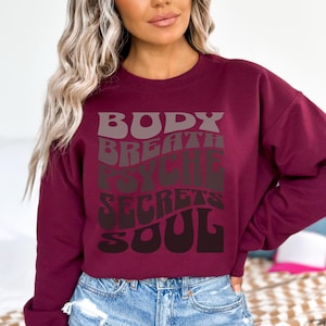 May include: Burgundy crewneck sweatshirt with the words "BODY, BREATH, PSYCHE, SECRETS, SOUL" in a layered, retro font. The text is in shades of gray and brown. The sweatshirt is a solid color and has a relaxed fit.
