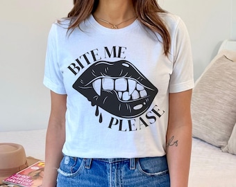 May include: White t-shirt with a black graphic of vampire lips and the text "BITE ME PLEASE". The design features sharp fangs and a drop of blood. The shirt is a casual, everyday wear item.