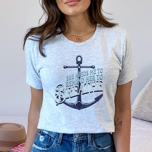 May include: A light gray t-shirt with a navy blue anchor graphic and the text "SHE NEEDS ME TO REMIND HER TO BREATHE" in blue and navy. The shirt is a crew neck style.