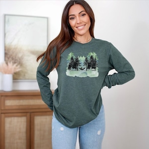 Op de afbeelding: Een donkergroen sweatshirt met lange mouwen en ronde hals, voorzien van een grafische print van zwarte dennenbomen en een persoon die ontspant in een hangmat, tegen een lichtgroene achtergrond in aquarel-stijl op de borst.