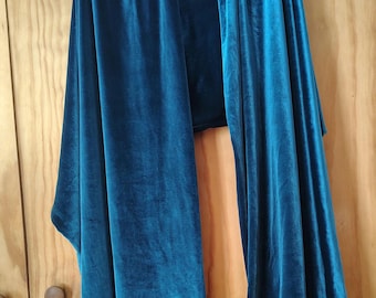 Könnte beinhalten: Ein langer, rechteckiger Schal aus blaugrünem Samt, der über einem dunklen Holzkleiderbügel hängt. Der Stoff hat den für Samt typischen weichen, reflektierenden Glanz mit tiefen blaugrünen Farbtönen und glatten, fließenden Falten.
