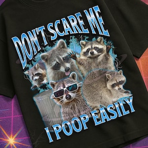 Op de afbeelding: Zwart T-shirt met een afbeelding van wasberen en de tekst "DON'T SCARE ME I POOP EASILY." Het shirt heeft ook de tekst "MILDLY PERTURBED" in een retro lettertype. De afbeelding heeft een vintage VHS-esthetiek.