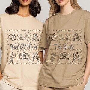 May include: Two women wearing beige t-shirts with matching graphic designs. The designs include wedding-themed illustrations and text that reads "Maid Of Honor" and "The Bride". The shirts have short sleeves and a relaxed fit.