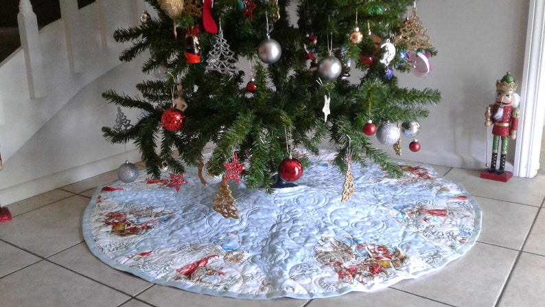 Puede incluir: Un &aacute;rbol de Navidad decorado con adornos y una falda de &aacute;rbol acolchada azul claro. La falda del &aacute;rbol presenta una escena invernal con ilustraciones rojas, blancas y marrones. Un cascanueces est&aacute; cerca.