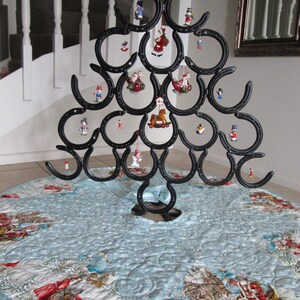Puede incluir: Un &aacute;rbol de Navidad de metal negro hecho de herraduras, decorado con adornos en miniatura. El &aacute;rbol se encuentra sobre una falda de &aacute;rbol acolchada azul claro con un dise&ntilde;o de escena invernal. El &aacute;rbol mide aproximadamente 91 cm de altura.