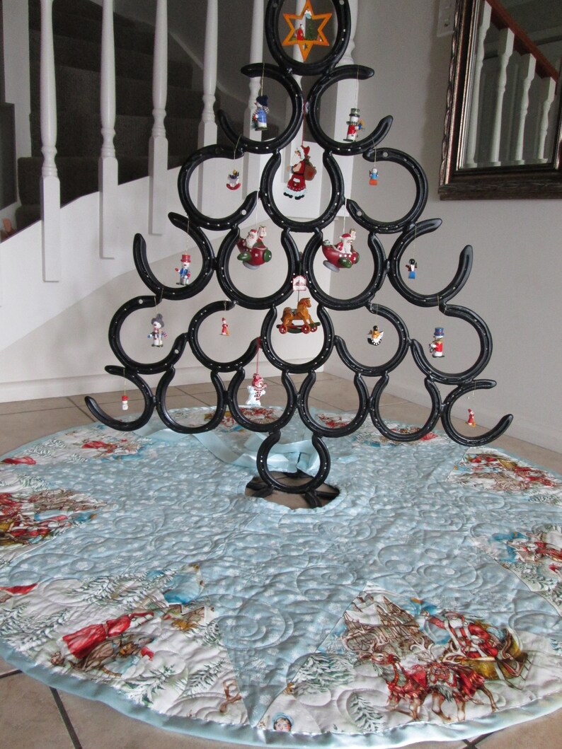 Puede incluir: Un &aacute;rbol de Navidad de metal negro hecho de herraduras, decorado con adornos en miniatura. El &aacute;rbol se encuentra sobre una falda de &aacute;rbol acolchada azul claro con un dise&ntilde;o de escena invernal. El &aacute;rbol mide aproximadamente 91 cm de altura.