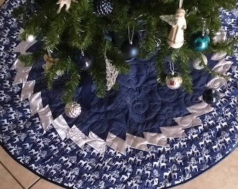 Puede incluir: Una falda de árbol de Navidad acolchada azul y blanca con ribete plateado y un patrón de renos blancos. La falda está decorada con adornos azules y plateados.