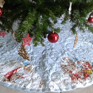 Puede incluir: Una falda redonda de &aacute;rbol de Navidad azul claro con un dise&ntilde;o acolchado. La falda presenta una escena invernal con figuras en rojo y blanco, copos de nieve y un borde azul claro. Adornos rojos y plateados cuelgan del &aacute;rbol.