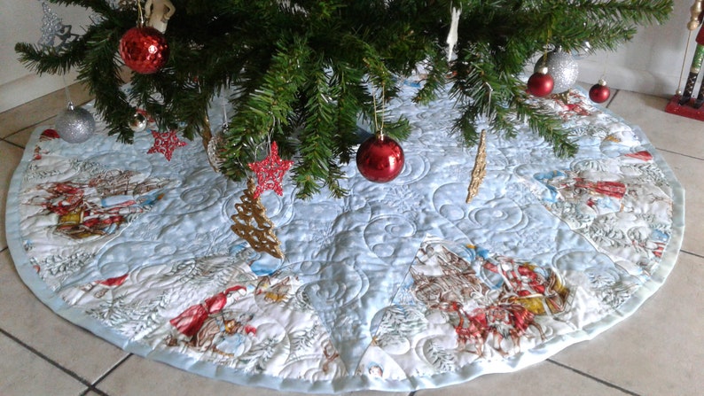 Puede incluir: Una falda redonda de &aacute;rbol de Navidad azul claro con un dise&ntilde;o acolchado. La falda presenta una escena invernal con figuras en rojo y blanco, copos de nieve y un borde azul claro. Adornos rojos y plateados cuelgan del &aacute;rbol.