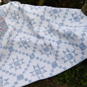 Puede incluir: Un edredón blanco y azul con un diseño de estrellas. El edredón presenta un diseño repetitivo de estrellas y cuadrados azules sobre un fondo blanco. El borde del edredón tiene un estampado floral en rosa, azul y blanco.