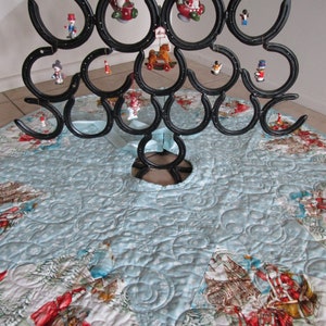Puede incluir: Un original &aacute;rbol de Navidad hecho de herraduras negras, adornado con adornos en miniatura. El &aacute;rbol se apoya en una falda de &aacute;rbol acolchada azul claro con un dise&ntilde;o de escena invernal. La falda tiene un borde de sat&eacute;n.