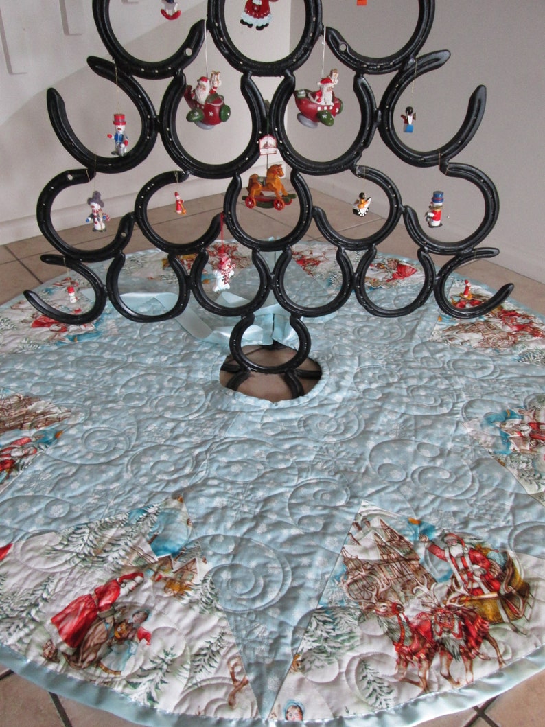 Puede incluir: Un original &aacute;rbol de Navidad hecho de herraduras negras, adornado con adornos en miniatura. El &aacute;rbol se apoya en una falda de &aacute;rbol acolchada azul claro con un dise&ntilde;o de escena invernal. La falda tiene un borde de sat&eacute;n.