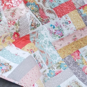 Puede incluir: Una colcha de retazos con un diseño floral y paisley. La colcha está hecha de cuadrados de diferentes telas, cada uno con un patrón único. La colcha tiene una paleta de colores pastel suave con tonos de rosa, azul, amarillo y verde.