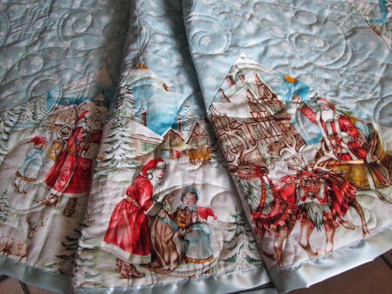 Puede incluir: Una falda de &aacute;rbol de Navidad acolchada de color azul claro con una escena invernal que presenta a Pap&aacute; Noel, renos y ni&ntilde;os. La falda tiene un borde de sat&eacute;n y un patr&oacute;n de copos de nieve.