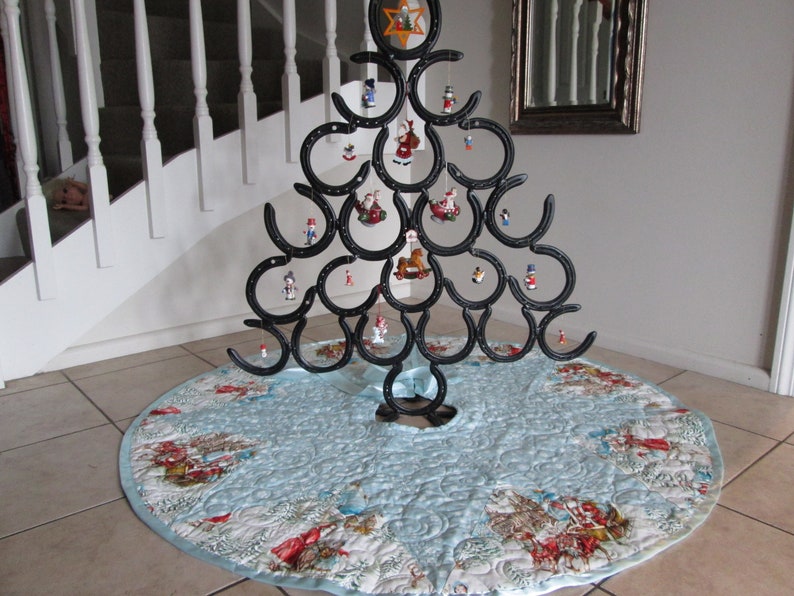 Puede incluir: Un original &aacute;rbol de Navidad hecho con herraduras negras, adornado con peque&ntilde;os adornos. El &aacute;rbol se encuentra sobre una falda de &aacute;rbol acolchada azul claro con una escena invernal. El &aacute;rbol mide aproximadamente 152 cm de alto.