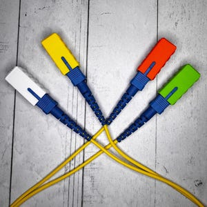 May include: Four fibre optic cables with coloured connectors: white, yellow, red, and green. The connectors are blue and the cables are yellow. The cables are arranged in a starburst pattern against a grey wooden background.