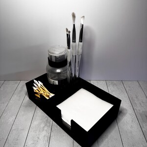 May include: A black artist's tray holds art supplies. The tray contains paint brushes, a jar, cotton swabs, and a stack of white paper. The brushes have white handles and silver ferrules. The jar is clear with a black band.