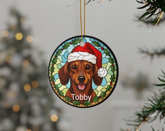 May include: Round Christmas ornament featuring a dachshund wearing a Santa hat. The dog is brown with a happy expression, set against a stained-glass style background with holly. The name "Tobby" is written below the dog.