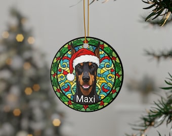 May include: Circular Christmas decoration with a Doberman Pinscher wearing a Santa hat. The dog is set against a stained-glass style background with holly and berries. The name "Maxi" is printed below the dog's face.
